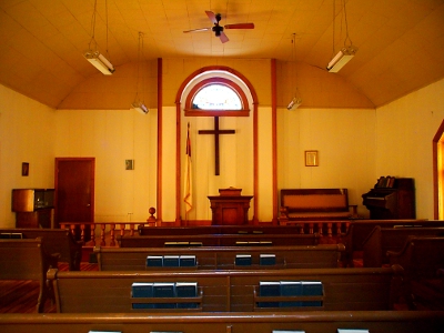 [Three columns of five dark brown pews leading up to a slightly raised pulpit with a cross hanging behind it and a curved window above the cross.]